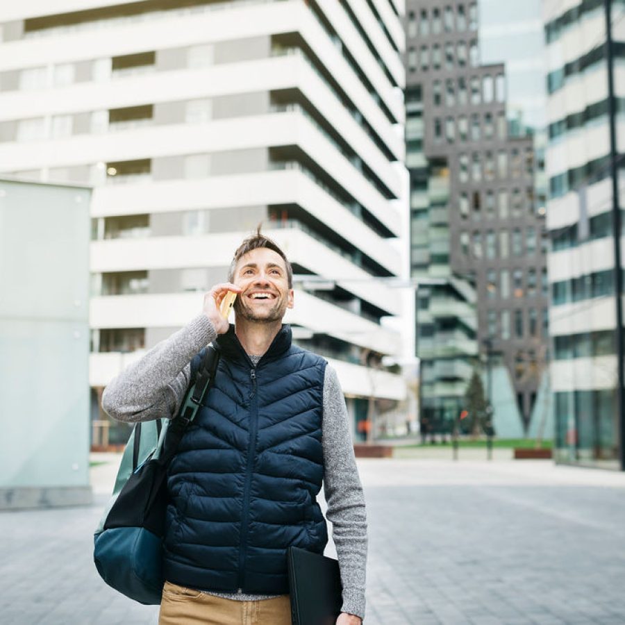 Freelancer male in casual clothes, using smartphone, modern building on background. Working remotely in urban location